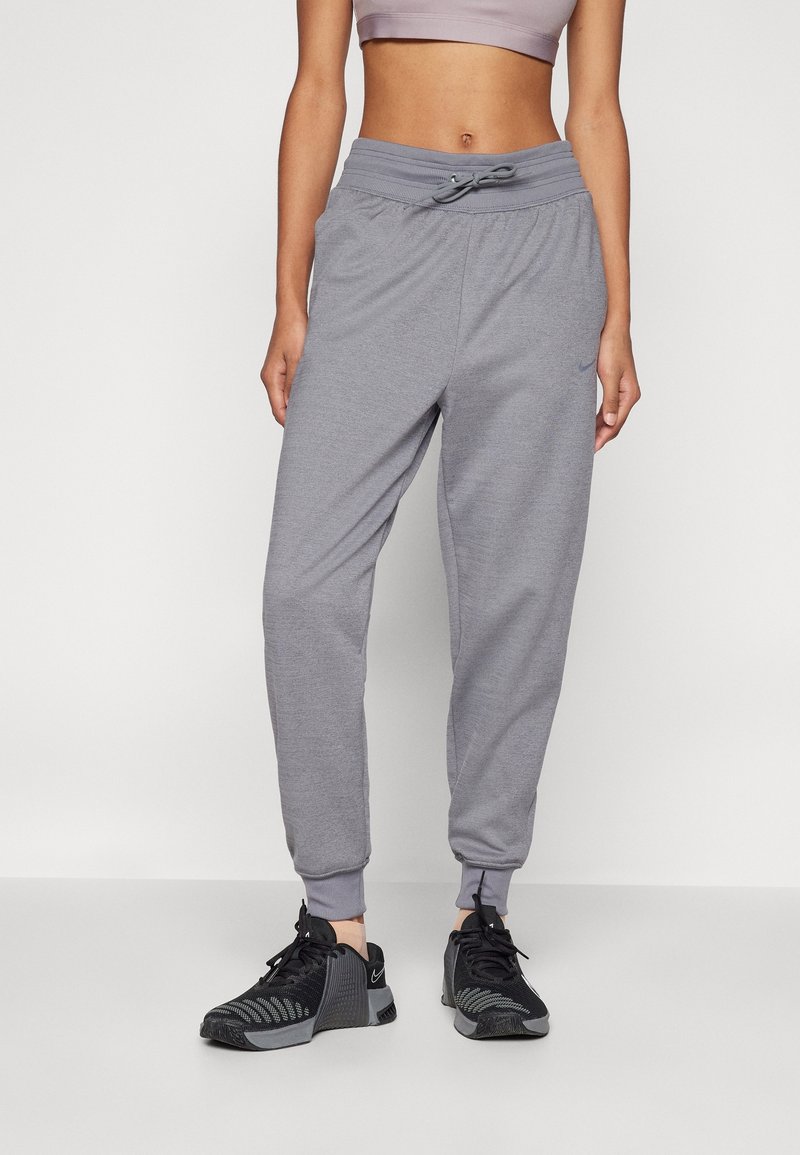 Femme portant un pantalon de jogging gris clair avec taille à cordon et des chaussures de sport noires, debout devant un fond clair uni.