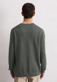 Mężczyzna odwrócony plecami, ubrany w sweter o fakturze w ciemnoszarym kolorze z długim rękawem oraz jasnobeżowe spodnie, stojący na tle gładkiej białej ściany.