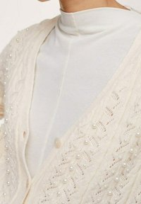 Kobieta w kremowym swetrze z dzianiny z perłowymi zdobieniami, nosząca biały top z wysokim kołnierzem, odsłaniająca górną część klatki piersiowej i ramienia.