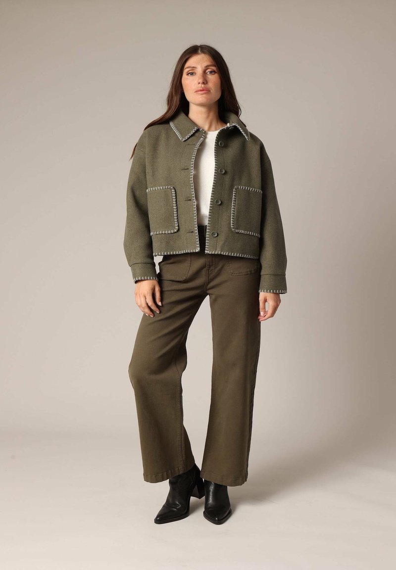 Veste courte vert olive avec des coutures blanches, grandes poches et fermeture à boutons. Associe à un pantalon vert olive et des bottines noires.