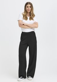 Pantalons larges noirs avec une texture lisse, une taille élastique et des poches latérales, associés à un t-shirt blanc ajusté et des baskets décontractées.