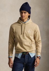 Bărbat tânăr purtând un hanorac Polo bej peste o cămașă albastră din denim, caciulă bleumarin, blugi și un ceas de mână, stând în fața unui fundal neutru.