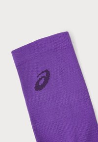 Lila sportstrumpa i texturerat tyg med en svart logotyp broderad nära den övre kanten. Enkel design utan ytterligare mönster.