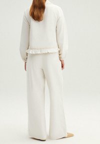 Mujer con cabello largo vestida con pantalones anchos blancos y una chaqueta a juego con ribetes ondulados, de pie contra un fondo claro y sencillo.
