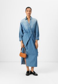 Lys blå denimskjortekjole med omslagdesign, lange ermer og sidespalter. Tilbehør med en liten brun håndveske og svarte loafers.