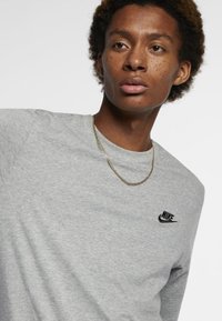 Camisola cinza de manga longa com o logótipo da Nike em preto, decote redondo e textura suave. Modelo a usar um colar fino em ouro.