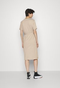 Vestido beige de manga curta com cinto na cintura, corte até ao joelho e fendas laterais, combinado com ténis pretos de cano alto. Textura de tecido suave.