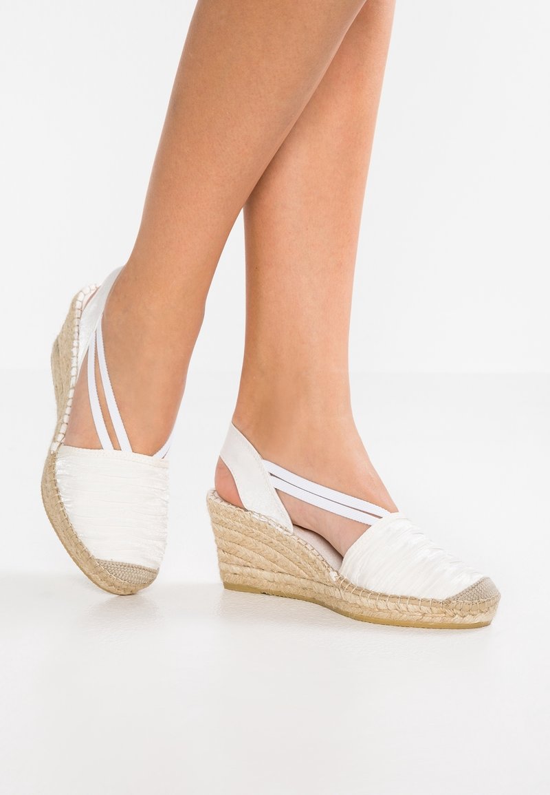 Jambes portant des espadrilles compensées blanches en tissu tissé avec semelles en jute et brides élastiques, debout sur une surface blanche.