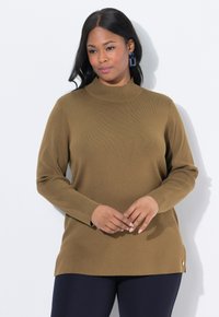 Frau in einem braunen Langarmpullover und blauen Hosen, die mit gefalteten Händen vor einem schlichten weißen Hintergrund steht.