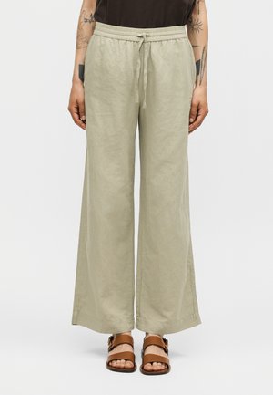 Osoba nosiaca voľné svetlo béžové ľanové nohavice so sťahovacou šnúrou v páse a hnedé sandále s otvorenou špičkou, stojaca pred jednofarebným bielym pozadím.