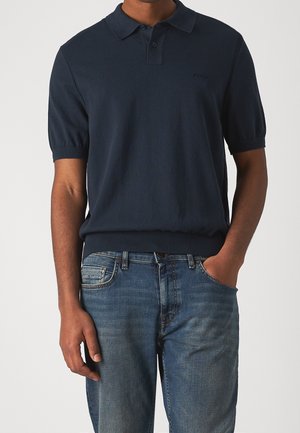 Polo en maille bleu marine à manches courtes, avec des poignets et un ourlet côtelés, présentant un détail de logo discret sur la poitrine. Associé à un jean en denim.