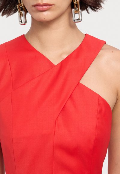 Robe sans manches rouge présentant un design de drapé diagonal avec une texture lisse, accompagnée de grandes boucles d'oreilles métalliques à clip.