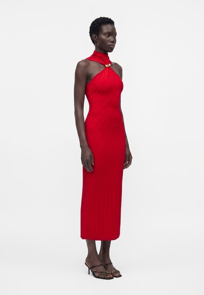 Γυναίκα με αμάνικο κόκκινο midi φόρεμα με halter neck και χρυσό κούμπωμα, στέκεται με μαύρα σανδάλια με λουράκια και τακούνι μπροστά σε απλό φόντο.