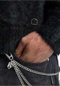Schwarzer, flauschiger Pullover mit einem kleinen Logodetail und einer Hand in der Tasche. Silberne Kettenaccessoires an dunklen Jeans.