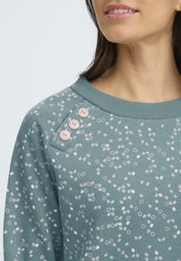 Felpa turchese con motivi circolari bianchi e rosa sparsi, con bottoni sulla spalla sinistra e un collo a coste. Tessuto morbido.