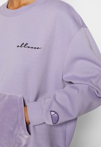 Sweat-shirt lavande avec un logo cursif noir sur la poitrine et un emblème semi-circulaire brodé sur la manche, main dans la poche avant.