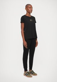 T-shirt de algodão preto com mangas curtas e um logótipo dourado, combinado com calças pretas e ténis verde-oliva com riscas azuis.