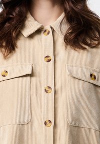 Beige väri beisvärinen corduroy-paita, jossa on nappikiinnitteinen kaulus, koristenappeja ja kaksi rintataskua. Kankaan pinnassa pystysuuntaiset uritukset.