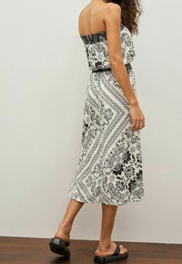 Frau in einem trägerlosen, schwarz-weiß geblümten Midikleid und schwarzen Riemchensandalen, die vor einer schlichten Wand steht.
