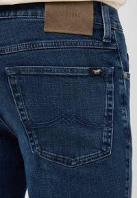 Ciemnoniebieskie dżinsy z denimu z naszywką "Mustang" ze skóry i charakterystycznym podwójnym przeszyciem na kieszeni. Tkanina o fakturze z gładkim wykończeniem.