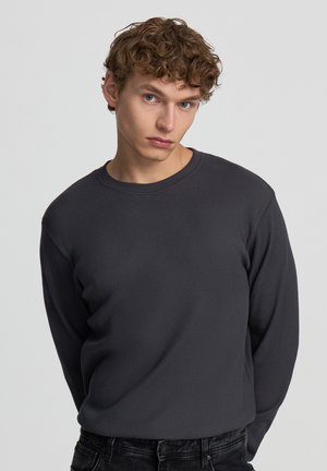 Maglietta a maniche lunghe grigia a coste con colletto rotondo, realizzata in tessuto testurizzato, abbinata a jeans neri. Il design è aderente, evidenziando la silhouette.