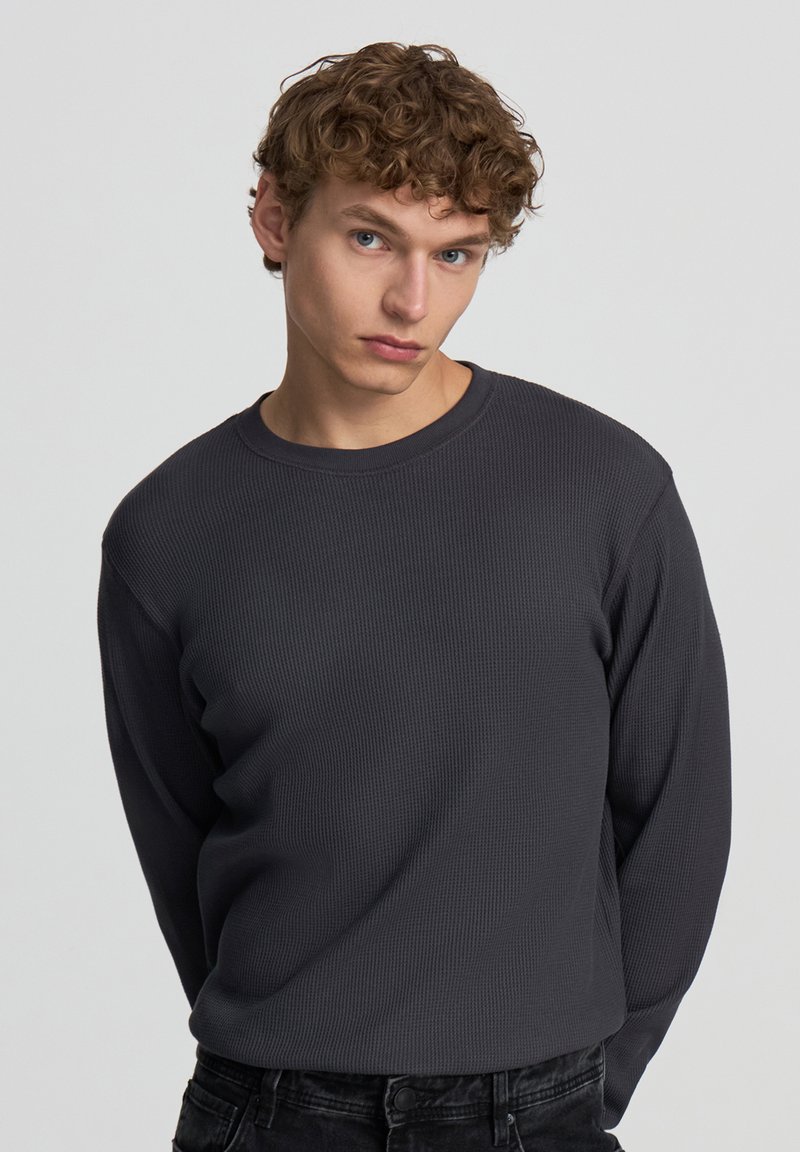 Szary, długi rękaw, prążkowany t-shirt z okrągłym dekoltem, wykonany z teksturowanej tkaniny, w połączeniu z czarnymi dżinsami. Krój jest dopasowany, podkreślający sylwetkę.