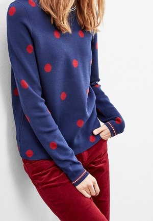 Maglione blu navy con grandi pois rossi, maniche lunghe e polsini a coste. Abbinato a pantaloni rossi con texture, dal taglio casual.