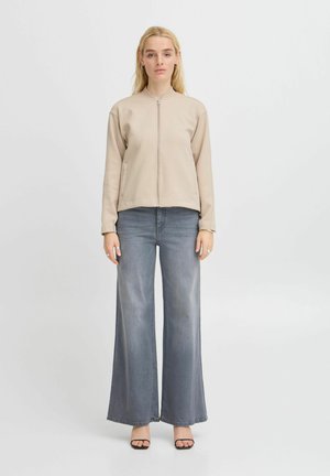 Beige Reißverschlussjacke mit Stehkragen, kombiniert mit hellgrauen, weiten Jeans, die eine glatte Textur und ein minimalistisches Design aufweisen.