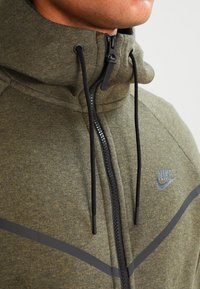 Olivgrön zip-up jacka med hög krage, svart dragkedja, snörning och en grå Nike-logga. Mjuk tyg med en slät textur.