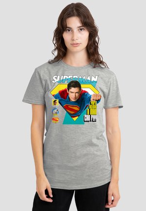 Woman wearing gray T-shirt featuring Superman flying with text "You'll believe a man can fly!" and small icons of Superman, dog, and logo.