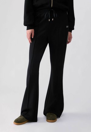 Personne portant un pantalon noir évasé avec taille à cordon et des baskets vertes, debout devant un fond uni clair.