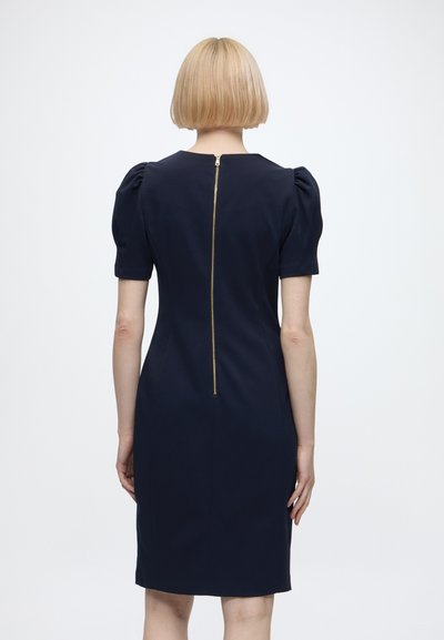 Femme aux cheveux blonds courts portant une robe bleu marine ajustée avec des manches bouffantes et une fermeture éclair dorée visible dans le dos.
