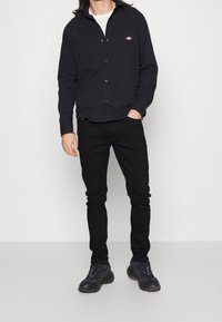 Man wearing black button-up shirt, black pants, and black sneakers standing with one hand in pocket against white background.