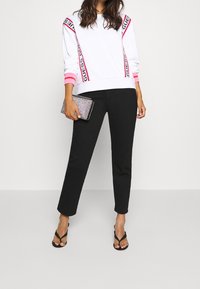 Mujer con sudadera blanca con franjas rosas, pantalones negros, sandalias negras, sosteniendo un bolso de mano brillante, de pie contra un fondo liso.