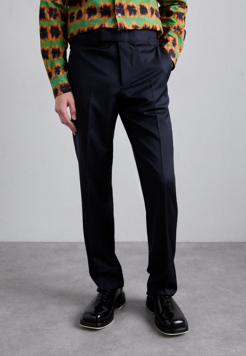 Pantalons ajustés bleu marine avec une texture lisse, dotés de poches latérales et d'un devant plat, associés à des chaussures noires à lacets.