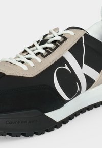 Μαύρο και μπεζ αθλητικό παπούτσι Calvin Klein με λευκά κορδόνια, μεγάλο λογότυπο "CK" στο πλάι και υφή στη μαύρη εξωτερική σόλα.
