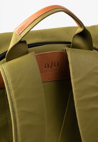 Borsa verde oliva con superficie in tessuto liscio, dotata di manico in pelle marrone e un'etichetta con logo impressa sulla tracolla interna.