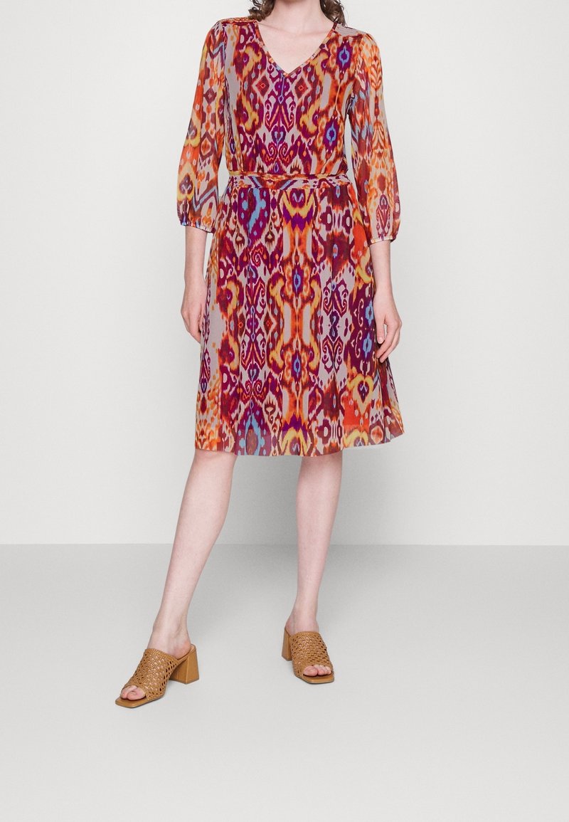 Robe multicolore avec un décolleté en V, des manches longues et un motif texturé orange, violet et rose. Associée à des sandales à talons bloc beiges.
