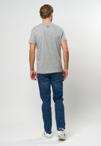 Graues Kurzarm-T-Shirt mit Rundhalsausschnitt, kombiniert mit dunkelblauen Jeans. Das T-Shirt hat ein dezentes Logodetail nahe des oberen Rückens.