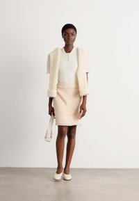 Ein kurzarmiger Jacke aus creamfarbigem Kunstfell über einem weißen gerippten Oberteil und einem strukturierten hellrosa, figurbetonten Rock. Das Model hält eine helle Tasche und trägt creamfarbene Schuhe.