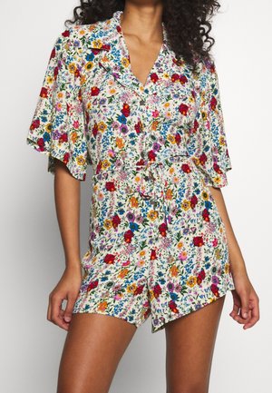 Donna che indossa un romper corto a fiori con maniche a svolazzo, bottoni frontali, colletto e vita con fiocco in fantasia floreale multicolore brillante.