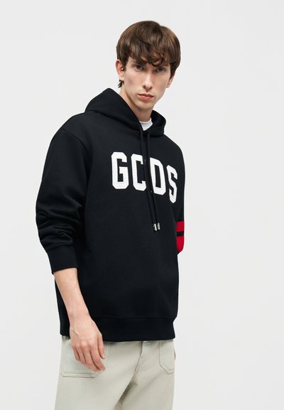 Ung mann iført svart hettejakke med bokstavene "GCDS" og beige bukser, stående foran en enkel lys grå bakgrunn.