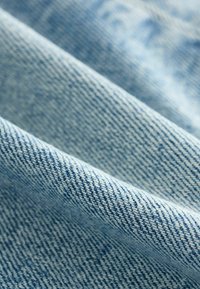 Tissu en denim bleu clair avec un tissage en sergé, présentant une texture visible et des lignes diagonales. Apparence douce et légèrement froissée.