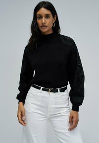 Pull noir à col haut avec des détails en dentelle florale sur les manches, associé à un pantalon blanc à jambes larges et une ceinture noire.