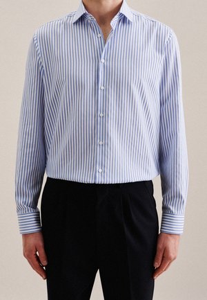 Un homme portant une chemise boutonnée à manches longues à rayures verticales bleues et blanches, rentrée dans un pantalon noir, debout devant un fond uni.