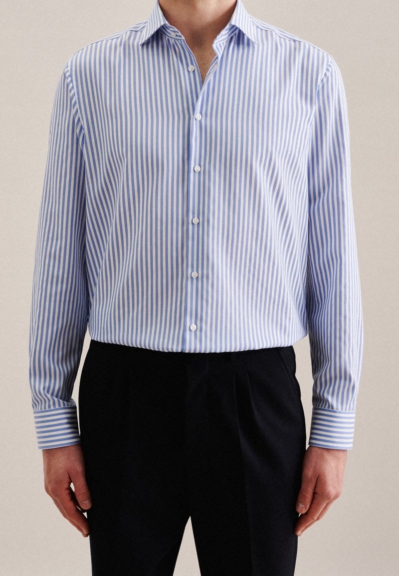 Un homme portant une chemise boutonnée à manches longues à rayures verticales bleues et blanches, rentrée dans un pantalon noir, debout devant un fond uni.