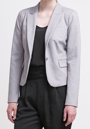 Nainen pukeutuneena istuvaan vaaleanharmaaseen bleiseriin mustan hihattoman topin ja mustien korkeavyötäröisten housujen päällä, seisoo yksinkertaisen taustan edessä.