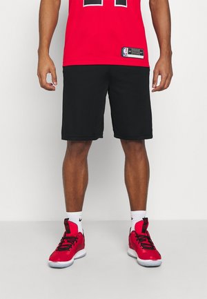 Mand stående iført sorte atletikshorts, rød basketballtrøje, hvide sokker og røde basketballsko mod en ensfarvet hvid baggrund.