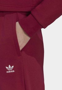 Burgundin värinen collegepaita, jossa on sivutasku, ribbineulehihansuut ja valkoinen brodeerattu Adidas-logo vasemmassa alakulmassa.