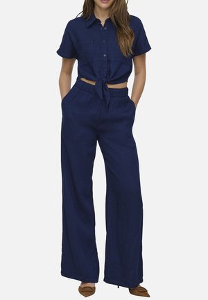 Donna che indossa una camicia blu navy a maniche corte con bottoni annodata in vita e pantaloni a gamba larga abbinati, con décolleté marroni a punta.